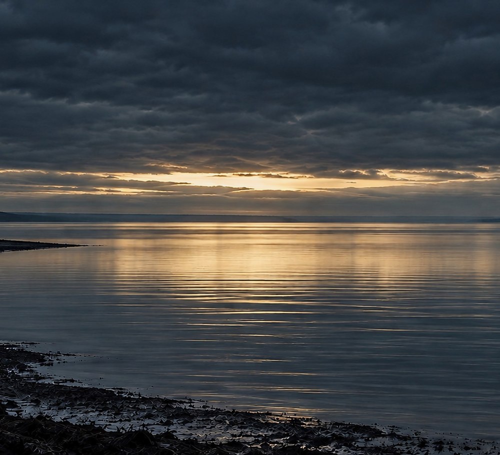 A wide, still bay at dusk under heavy cloud, with one long break of pale light along the horizon and a dark curve of shoreline at the left